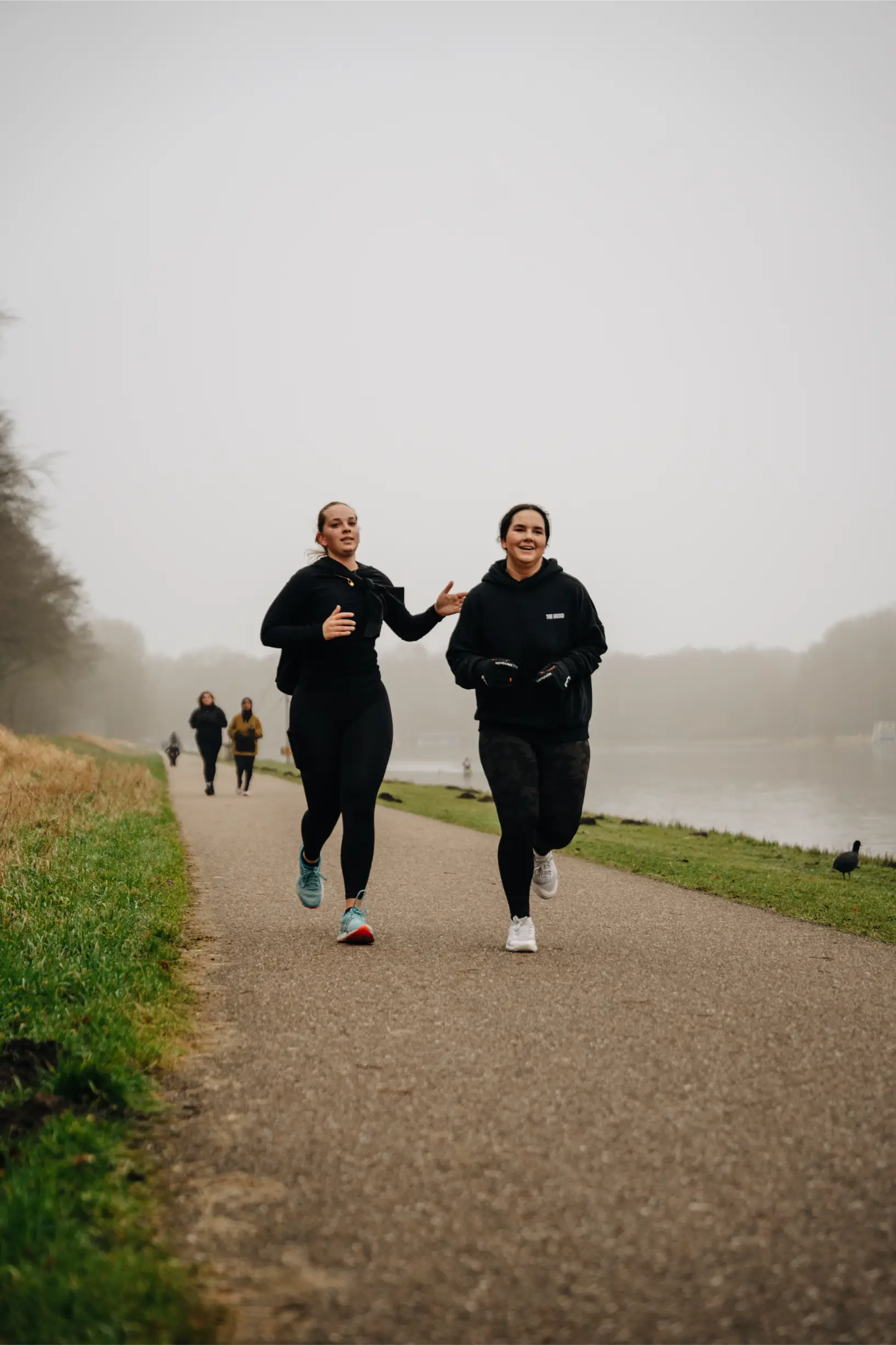 Zwei Frauen laufen an einem nebligen Tag an einem Fluss entlang, gekleidet in Sportbekleidung.