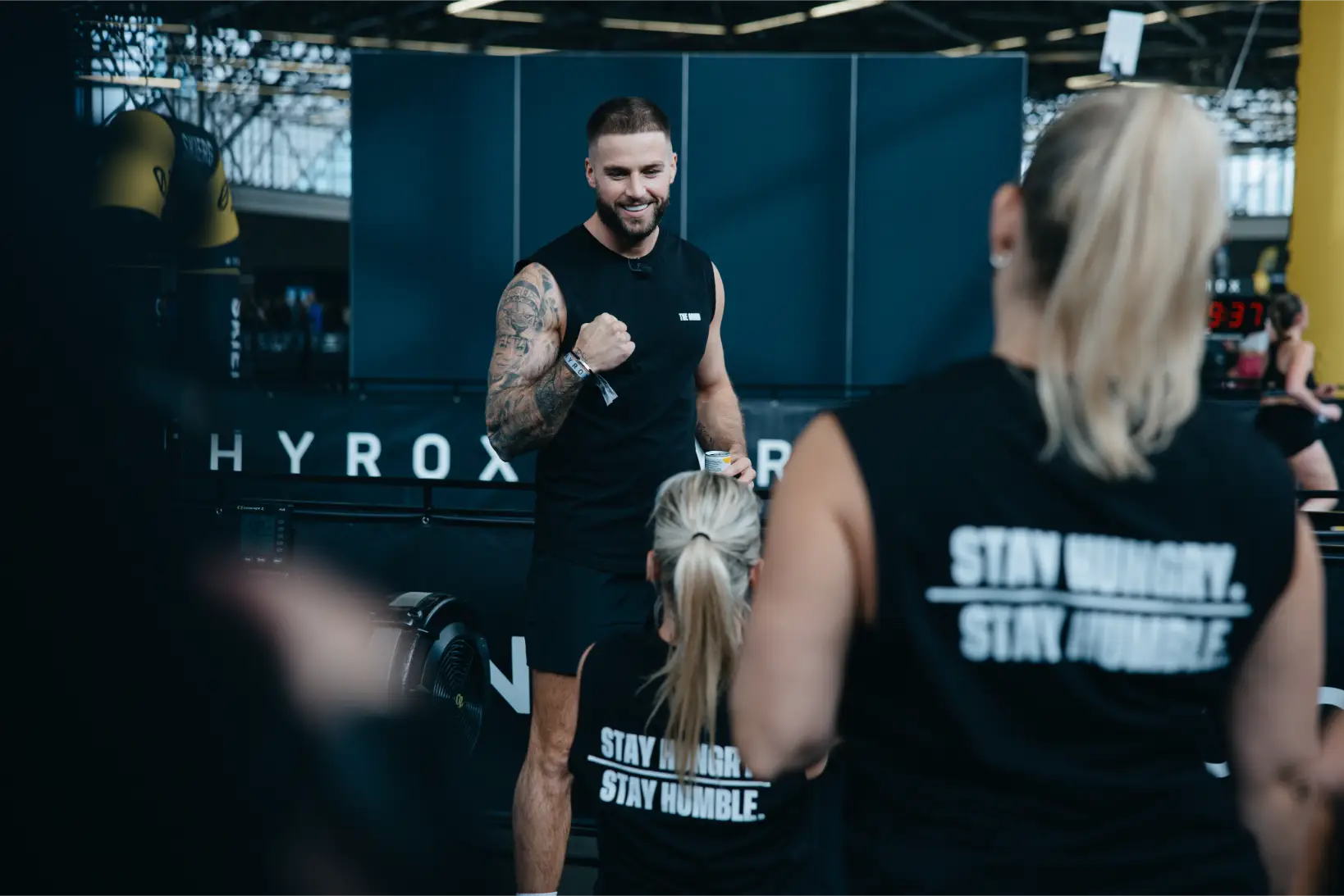 Ein tätowierter Mann ermutigt zwei Frauen, die schwarze T-Shirts mit der Aufschrift 'Stay Hungry, Stay Humble' in einem Fitnessstudio tragen.