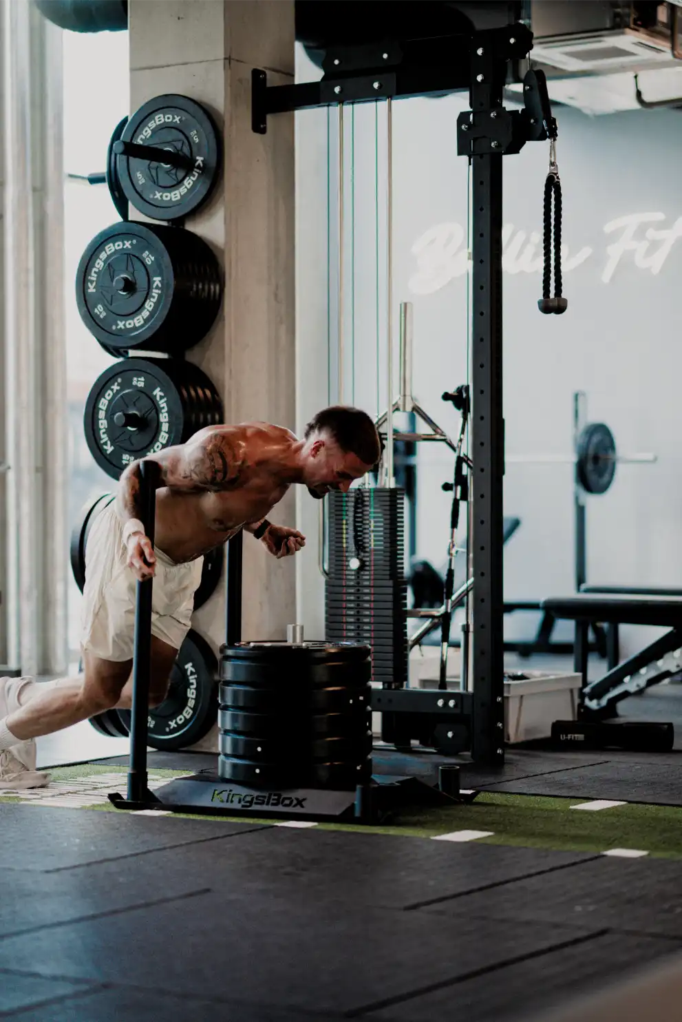 Muskulöser Mann führt eine Push-Sled-Übung in einem Fitnessstudio aus, umgeben von Gewichtsscheiben und Fitnessgeräten.