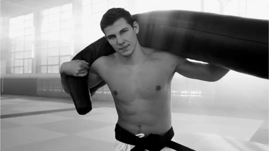 Junger Mann mit schwarzem Band und ohne Oberkörper, der einen schweren Boxsack auf den Schultern in einem beleuchteten Fitnessstudio trägt.