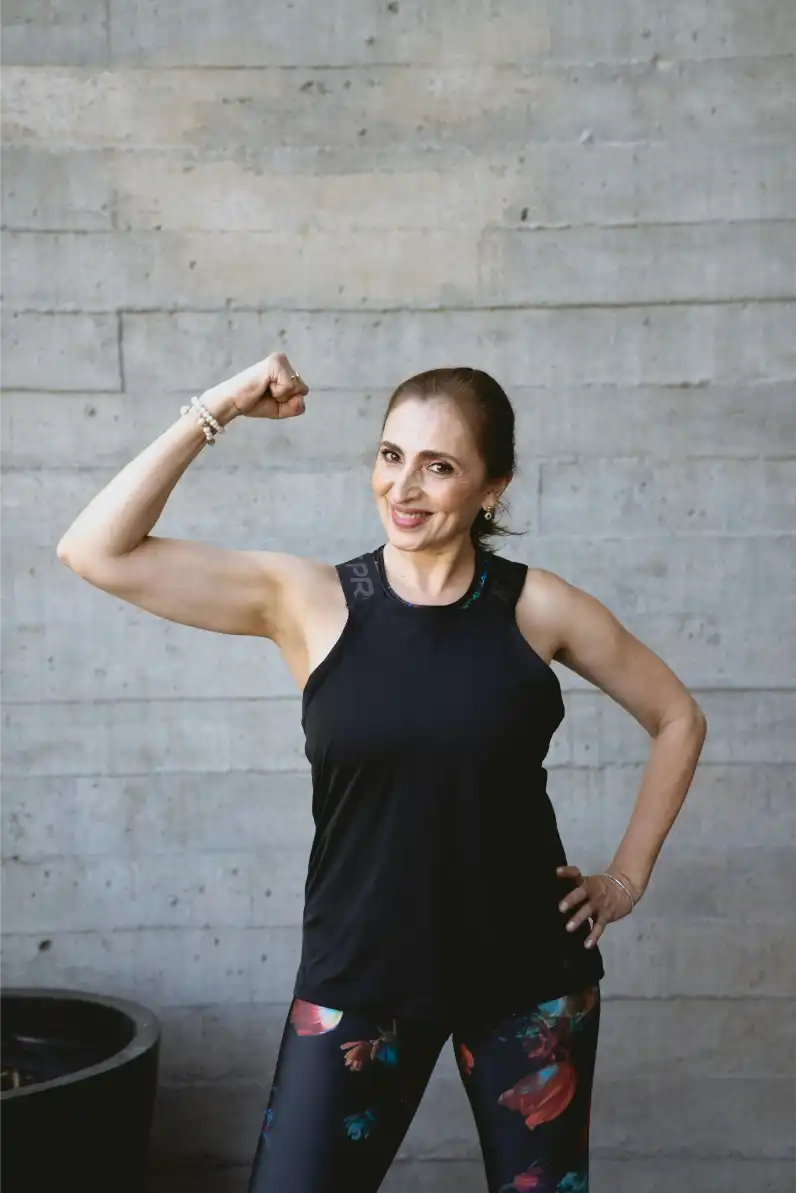 Frau in Sportbekleidung, die lächelt und ihre Bizeps vor einer Betonwand zeigt.